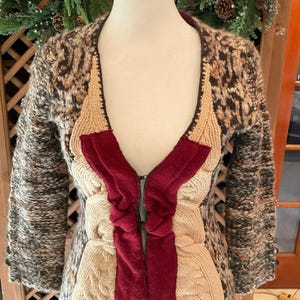 May include: A unique, handmade cardigan with a mix of textures and colors. The jacket features a patterned, speckled outer layer, with cream-colored cable knit and a deep red velvet-like fabric down the front. The cardigan has a zipper closure.