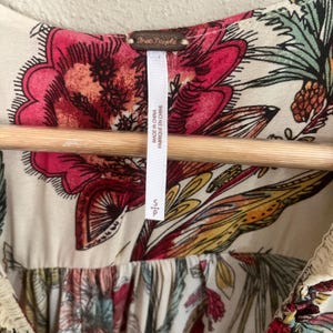May include: Cream-coloured top with a vibrant floral pattern in shades of pink, red, yellow, and green. The top features a scoop neckline and a tag that reads "Free People" and "Made in China". The size is small.
