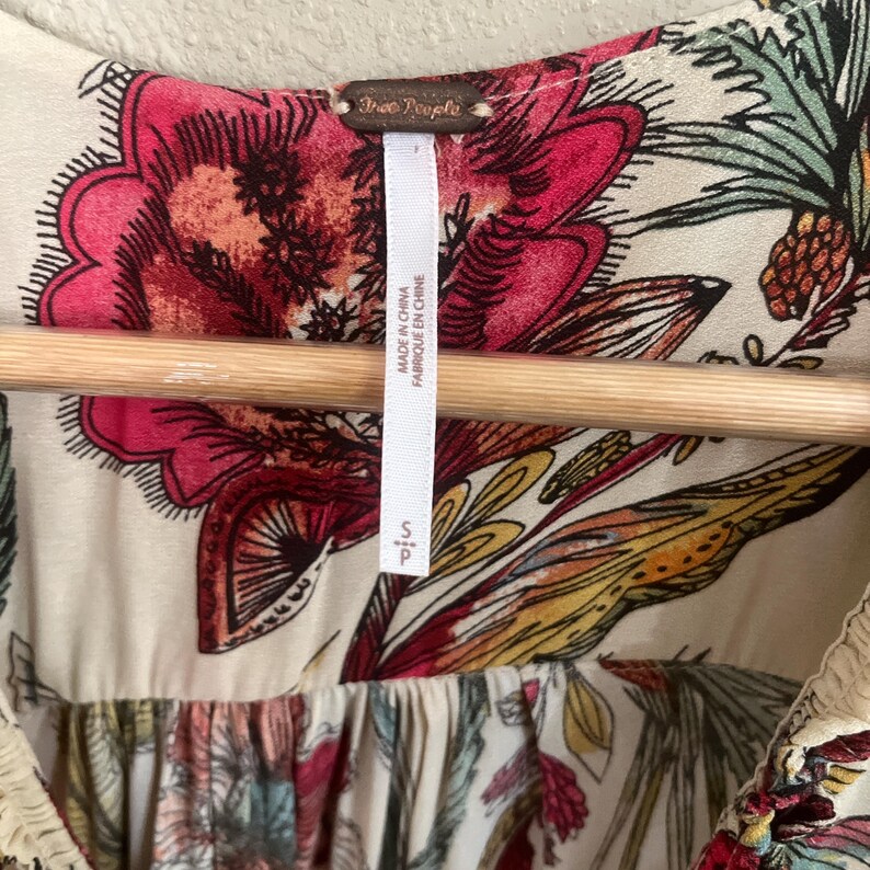 May include: Cream-coloured top with a vibrant floral pattern in shades of pink, red, yellow, and green. The top features a scoop neckline and a tag that reads "Free People" and "Made in China". The size is small.