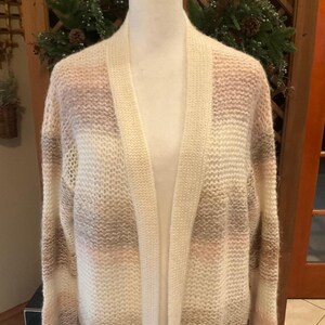 May include: A cream-colored open-front cardigan sweater with horizontal stripes of light brown, pink, and white. The sweater is displayed on a mannequin and has long sleeves. The knitwear is soft and cozy, perfect for layering.