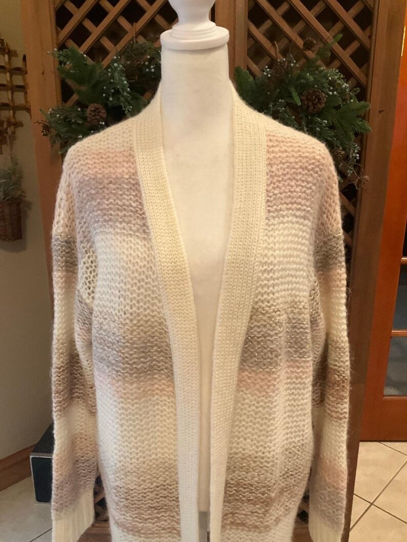 May include: A cream-colored open-front cardigan sweater with horizontal stripes of light brown, pink, and white. The sweater is displayed on a mannequin and has long sleeves. The knitwear is soft and cozy, perfect for layering.