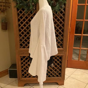 May include: A white, long-sleeved sweater with flared sleeves. The sweater is made of a thick knit material and has a V-neck. The sweater is displayed on a mannequin.