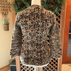 May include: A textured, multi-colored knit jacket with a round neckline and three-quarter sleeves. The jacket features a pattern of brown, beige, and black yarns. The sleeves have three dark buttons. The jacket is displayed on a mannequin.