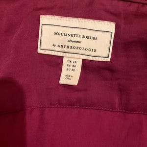 Puede incluir: Primer plano de una etiqueta de prenda. La etiqueta blanca presenta texto negro: "MOULINETTE SOEURS v&ecirc;tements by ANTHROPOLOGIE." Debajo, una etiqueta m&aacute;s peque&ntilde;a muestra las tallas: UK 10, US 06, EU 38. La prenda es de color magenta oscuro.