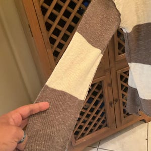 May include: A long-sleeved sweater with a color block design. The sleeve features alternating sections of brown and cream colors. The sweater is made of a soft, textured material.