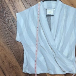 May include: A light gray and white striped wrap top with short sleeves. A measuring tape is placed vertically along the garment. The top has a cross-over design and a small label is visible.