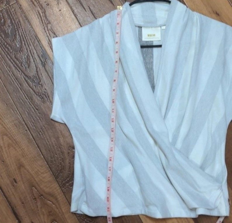 May include: A light gray and white striped wrap top with short sleeves. A measuring tape is placed vertically along the garment. The top has a cross-over design and a small label is visible.