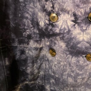 May include: A close-up of a tie-dye patterned jacket in shades of blue and white. The jacket features large, round, brown buttons and a front pocket. The fabric has a textured appearance.
