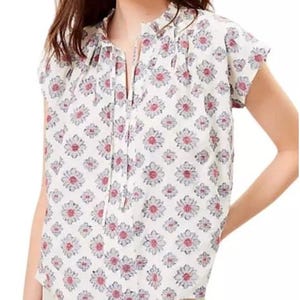 May include: White floral print top with short sleeves and a drawstring neckline. The top features a pattern of pink and gray flowers. The model is wearing beige shorts. This top is perfect for spring and summer.