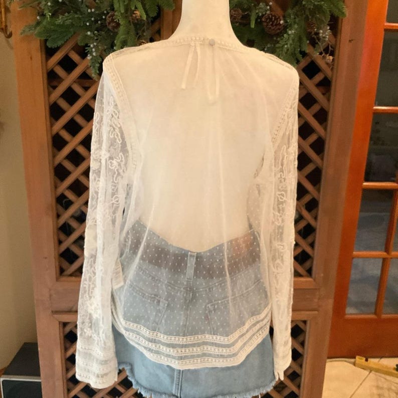 Pu&ograve; includere: Un top trasparente color crema a maniche lunghe con intricati dettagli in pizzo sulle maniche e sullo scollo. Il top &egrave; abbinato a una gonna di jeans lavata chiara. La parte posteriore del top &egrave; visibile, mostrando il tessuto e il design delicati.