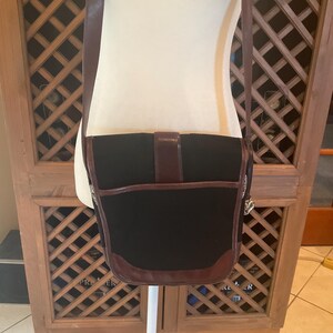 May include: Black satchel bag with brown leather trim and shoulder strap. The bag has a flap closure with a buckle. The bag is displayed on a white mannequin.