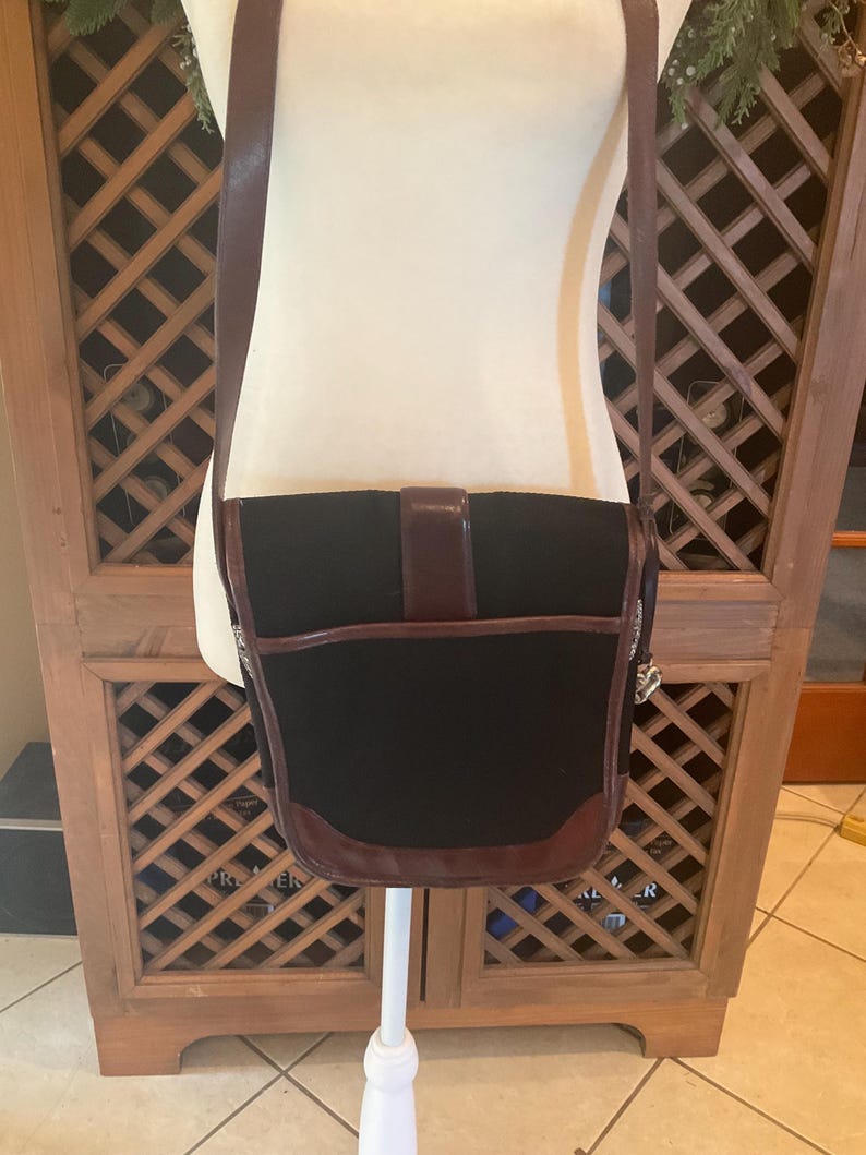 May include: Black satchel bag with brown leather trim and shoulder strap. The bag has a flap closure with a buckle. The bag is displayed on a white mannequin.