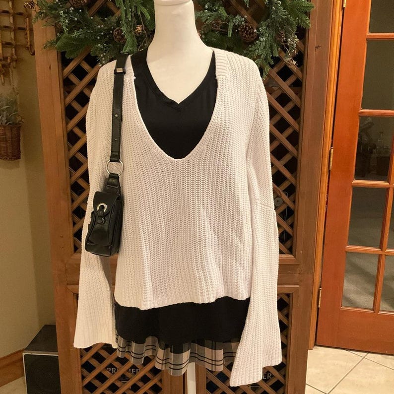 May include: A white, oversized sweater with a deep V-neck, layered over a black top. A small black purse with a shoulder strap hangs on the left. The bottom layer is a black and plaid skirt. The outfit is displayed on a mannequin.