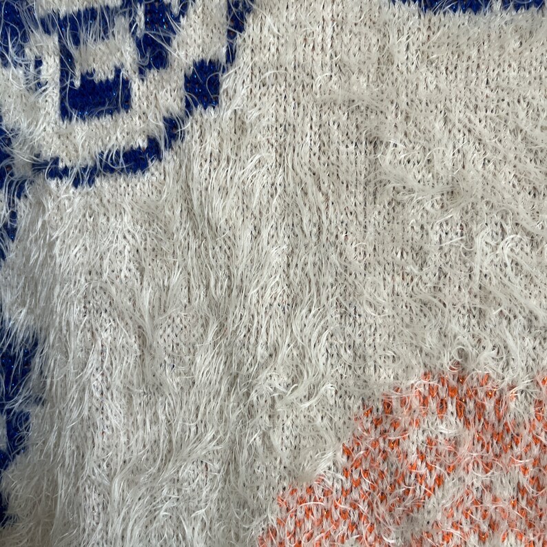 May include: A white fuzzy knit fabric with blue and orange geometric patterns. The fabric is soft and fluffy, and the patterns are intricate.