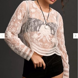 Pu&ograve; includere: Un top in pizzo bianco a maniche lunghe, con motivi floreali e a pois. Il top ha uno scollo tondo e viene indossato con pantaloncini neri. &Egrave; visibile anche una collana nera con un ciondolo bianco.