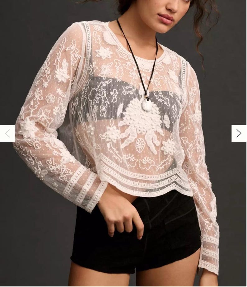 Pu&ograve; includere: Un top in pizzo bianco a maniche lunghe, con motivi floreali e a pois. Il top ha uno scollo tondo e viene indossato con pantaloncini neri. &Egrave; visibile anche una collana nera con un ciondolo bianco.