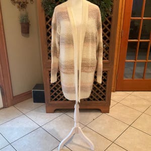 May include: A long, open-front cardigan in a neutral color palette of cream, tan, and blush pink stripes. The sweater is displayed on a white mannequin, showcasing its relaxed fit and open design. The knitwear is a cozy, stylish layering piece.