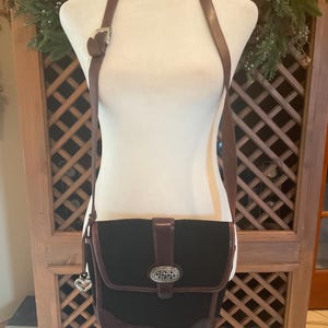 May include: Black crossbody bag with brown leather trim and strap. The bag features a decorative silver-toned buckle and a heart-shaped charm. The bag is displayed on a mannequin.