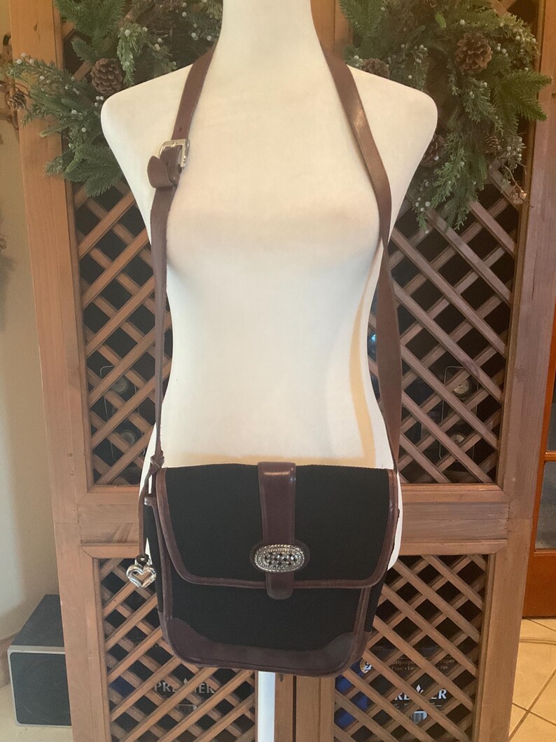 May include: Black crossbody bag with brown leather trim and strap. The bag features a decorative silver-toned buckle and a heart-shaped charm. The bag is displayed on a mannequin.