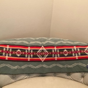 May include: A sage green wool blanket featuring a central horizontal band with a geometric pattern in red, orange, yellow, purple, and white. The blanket has a dark stitched edge and a small rectangular label in the bottom right corner.