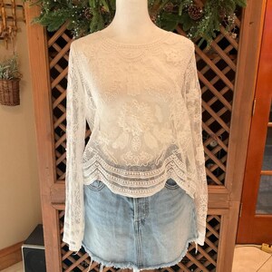 Pu&ograve; includere: Un top in pizzo color crema a maniche lunghe, con intricati motivi floreali e geometrici. &Egrave; abbinato a una gonna di jeans lavata chiara. Il pizzo offre un aspetto delicato e femminile. I motivi intricati sono visibili.