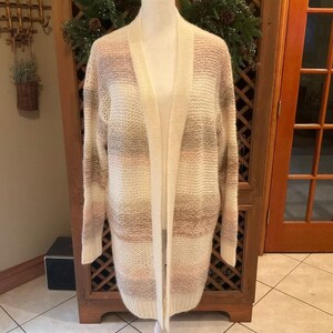 May include: A long, open-front cardigan in a neutral color palette of cream, pink, and tan stripes. The sweater is displayed on a white mannequin, showcasing its length and relaxed fit. The cardigan has long sleeves and two front pockets.