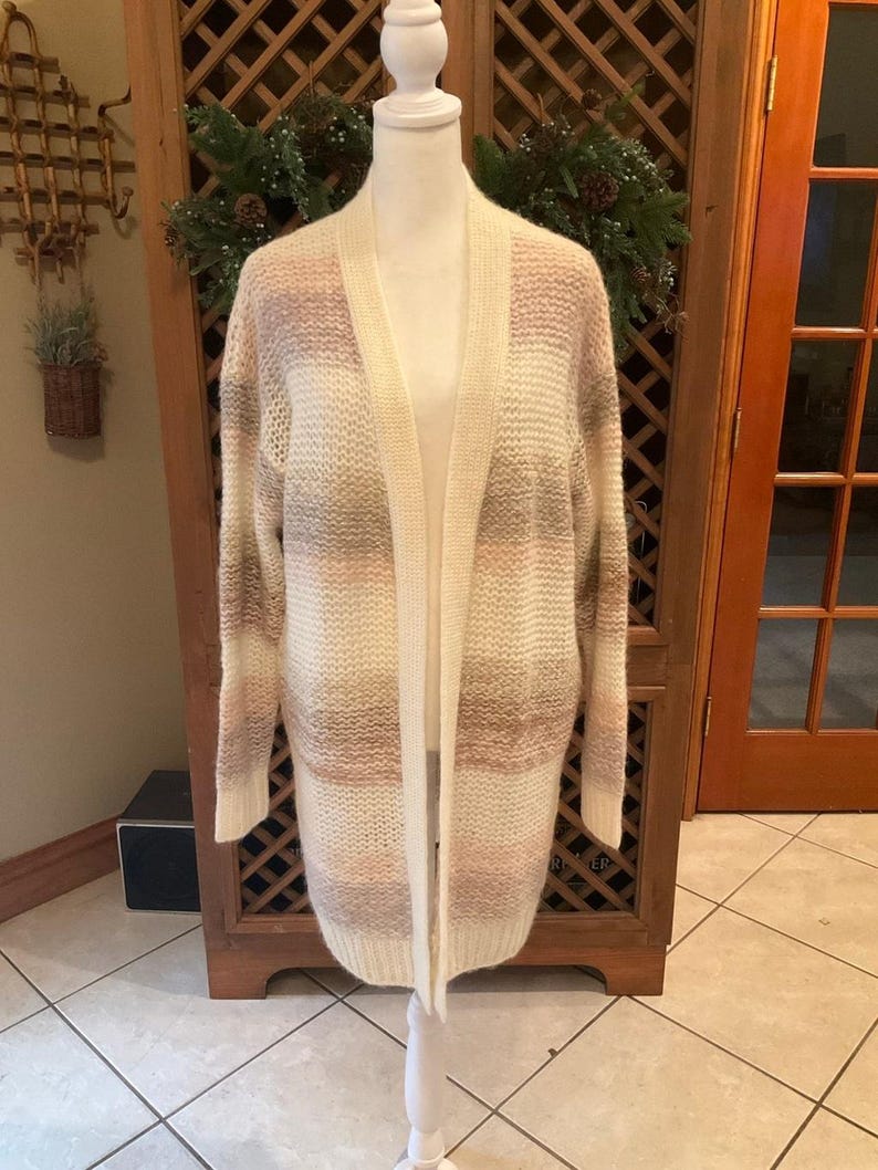 May include: A long, open-front cardigan in a neutral color palette of cream, pink, and tan stripes. The sweater is displayed on a white mannequin, showcasing its length and relaxed fit. The cardigan has long sleeves and two front pockets.