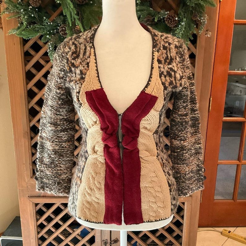 May include: A unique, textured cardigan with a mix of patterns and colors. The sleeves and upper body feature a speckled brown and tan knit, while the front showcases a combination of cream-colored cable knit and burgundy panels. It has a zipper closure.