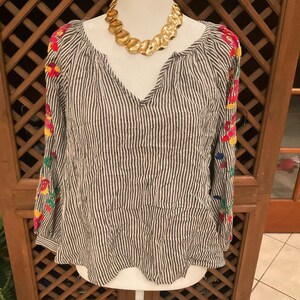 May include: A striped, long-sleeved blouse with a V-neck and colourful floral embroidery on the sleeves. A gold-toned statement necklace is draped around the neckline. The blouse is displayed on a white mannequin.