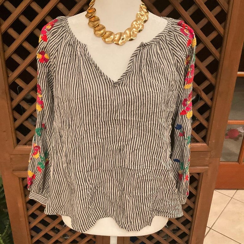 May include: A striped, long-sleeved blouse with a V-neck and colourful floral embroidery on the sleeves. A gold-toned statement necklace is draped around the neckline. The blouse is displayed on a white mannequin.
