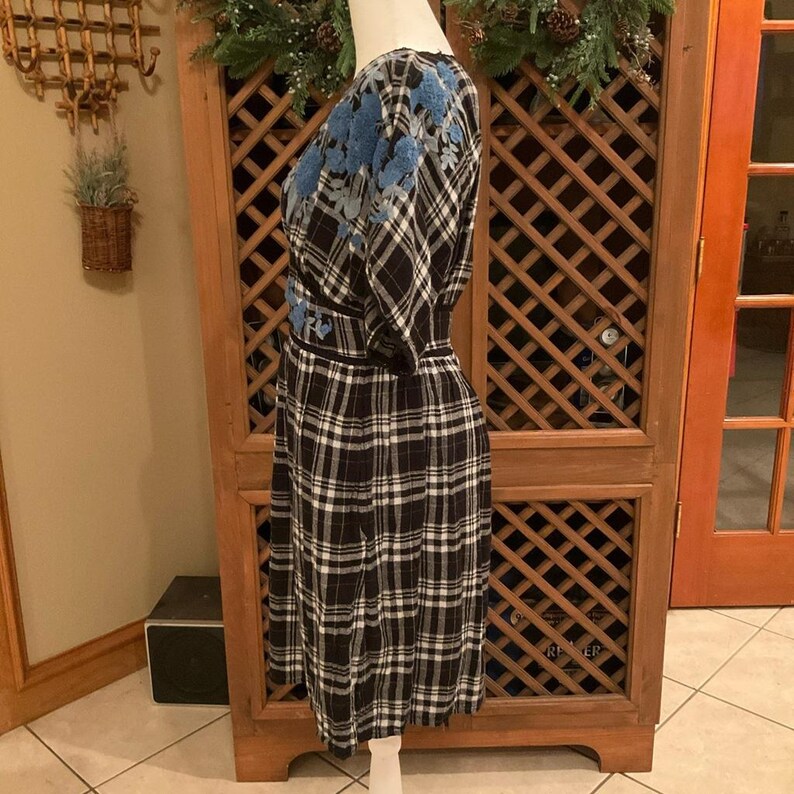 May include: A black and white plaid dress with blue floral embroidery on the shoulders and waist. The dress has a wrap-style bodice and a fitted waist. The dress is displayed on a mannequin.