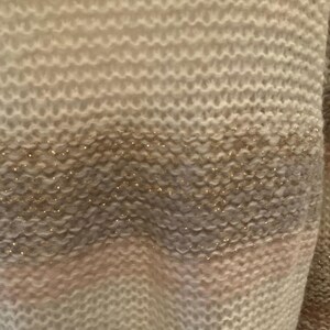 May include: Close-up of a knitted sweater with horizontal stripes in shades of cream, gray, and pink. The sweater has a textured pattern with subtle gold glitter accents, creating a soft and cozy appearance.