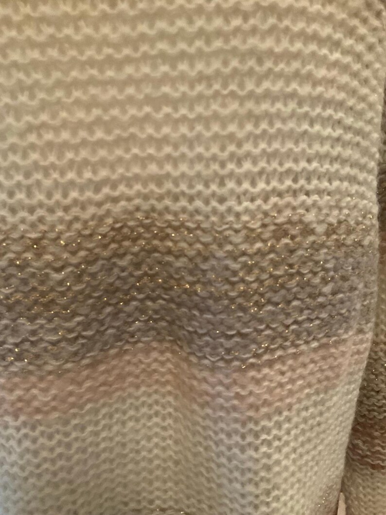 May include: Close-up of a knitted sweater with horizontal stripes in shades of cream, gray, and pink. The sweater has a textured pattern with subtle gold glitter accents, creating a soft and cozy appearance.