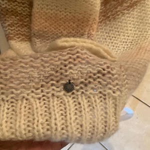 May include: A beige and light brown knitted sweater with a ribbed cuff and a small, decorative metal emblem. The sweater has a gradient effect, transitioning from light to darker shades of brown.