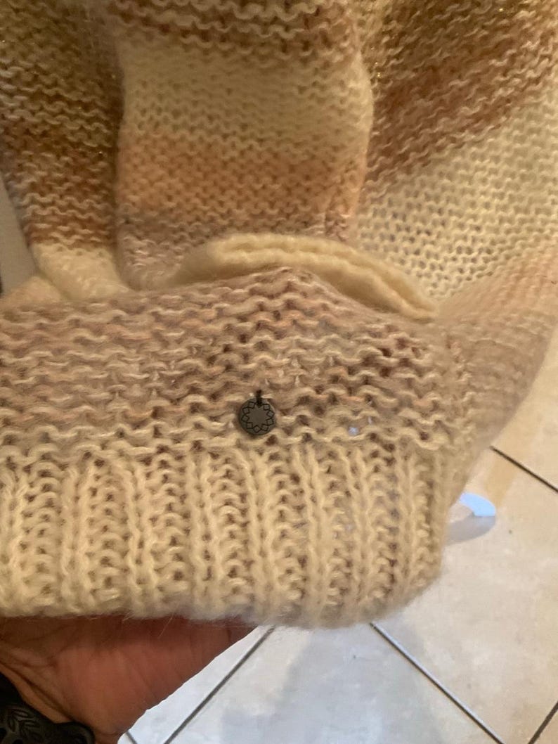 May include: A beige and light brown knitted sweater with a ribbed cuff and a small, decorative metal emblem. The sweater has a gradient effect, transitioning from light to darker shades of brown.