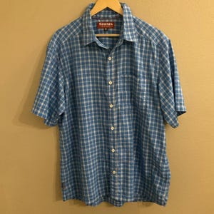 May include: A short-sleeved, button-down shirt in a blue and white plaid pattern. The shirt has a collar and a chest pocket. The brand name "SIMMS" is visible on a red tag near the collar. The shirt is hanging on a wooden hanger.