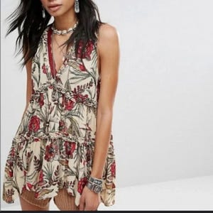 May include: Cream-coloured sleeveless top with a floral pattern in red and green. The top has a V-neckline and ruffled detailing. The fabric appears lightweight and flowy, suitable for warm weather.