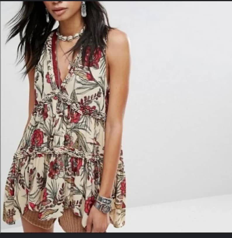 May include: Cream-coloured sleeveless top with a floral pattern in red and green. The top has a V-neckline and ruffled detailing. The fabric appears lightweight and flowy, suitable for warm weather.