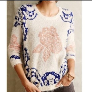 May include: A white fuzzy sweater with a pink rose design and blue and white geometric patterns around the edges.