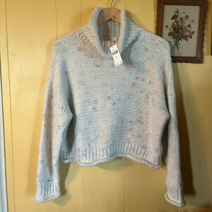 May include: A white, cropped, turtleneck sweater with a speckled pattern of pink, blue, and green. The sweater has long sleeves and a ribbed hem.
