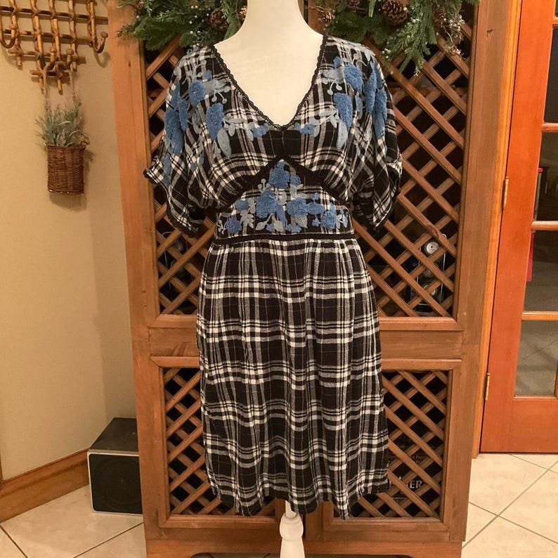 May include: A black and white plaid dress with blue floral embroidery on the shoulders and waist. The dress has a V-neck and short sleeves. The dress is displayed on a mannequin.