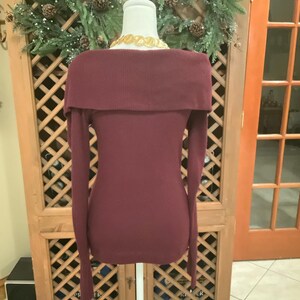Puede incluir: Un su&eacute;ter burdeos con hombros descubiertos y mangas largas exhibido en un maniqu&iacute;. El su&eacute;ter tiene una textura acanalada y un cuello ancho. Un collar dorado es visible en el escote. El fondo incluye un armario de madera y una corona navide&ntilde;a.