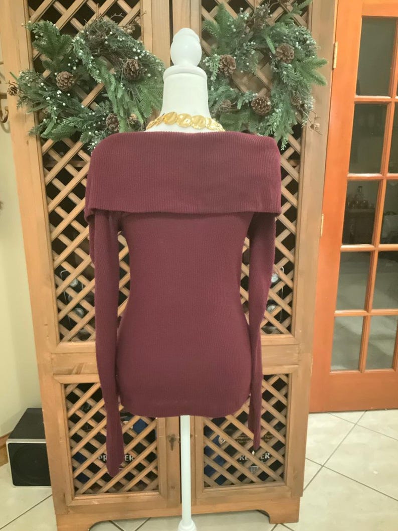 Puede incluir: Un su&eacute;ter burdeos con hombros descubiertos y mangas largas exhibido en un maniqu&iacute;. El su&eacute;ter tiene una textura acanalada y un cuello ancho. Un collar dorado es visible en el escote. El fondo incluye un armario de madera y una corona navide&ntilde;a.