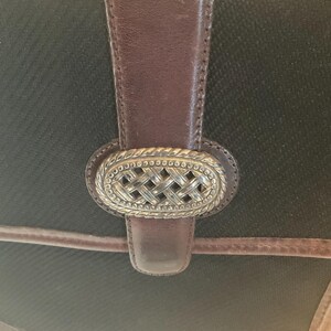 May include: Close-up of a black handbag with brown leather accents. The bag features a decorative silver-colored clasp with a woven design. The leather strap and trim add a classic touch to the bag's design.