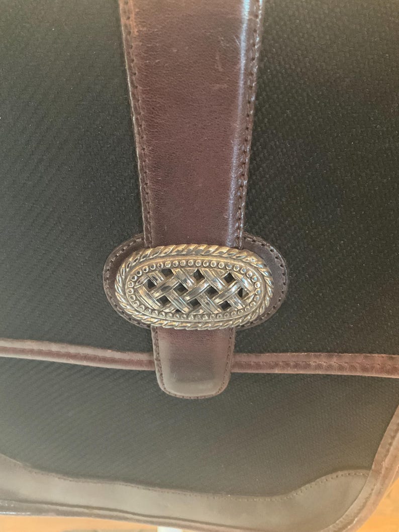 May include: Close-up of a black handbag with brown leather accents. The bag features a decorative silver-colored clasp with a woven design. The leather strap and trim add a classic touch to the bag's design.
