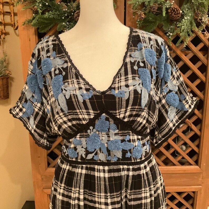 May include: A black and white plaid dress with a V-neckline and short sleeves. The dress features blue floral embroidery on the bodice and sleeves, with black lace trim. The dress is displayed on a mannequin.