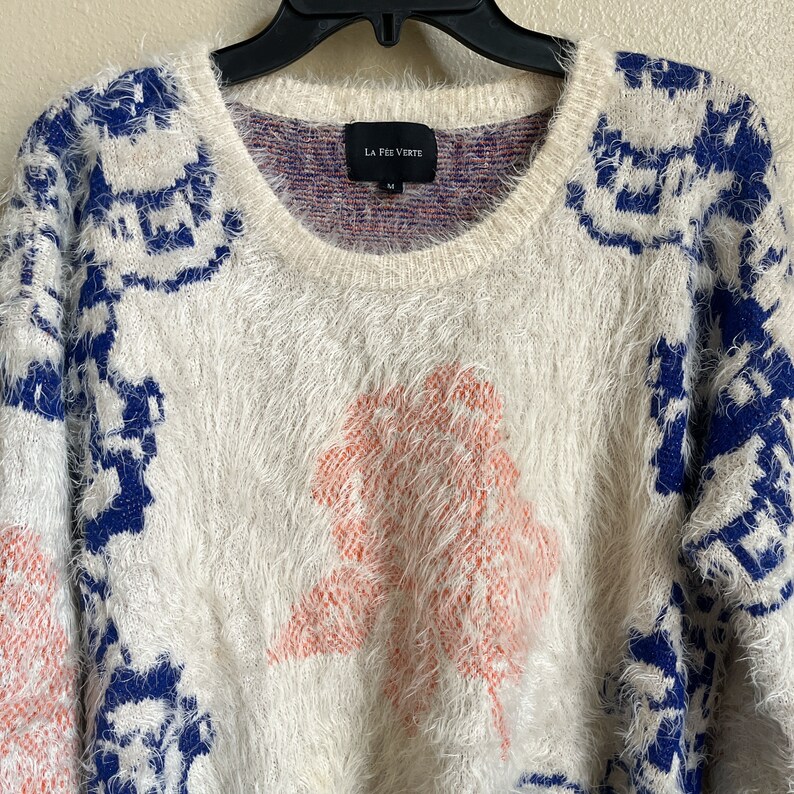 May include: White fuzzy sweater with a blue and white geometric pattern and a pink fuzzy tree design. The sweater has a crew neck and the brand label "La Free Verte" is visible on the front.