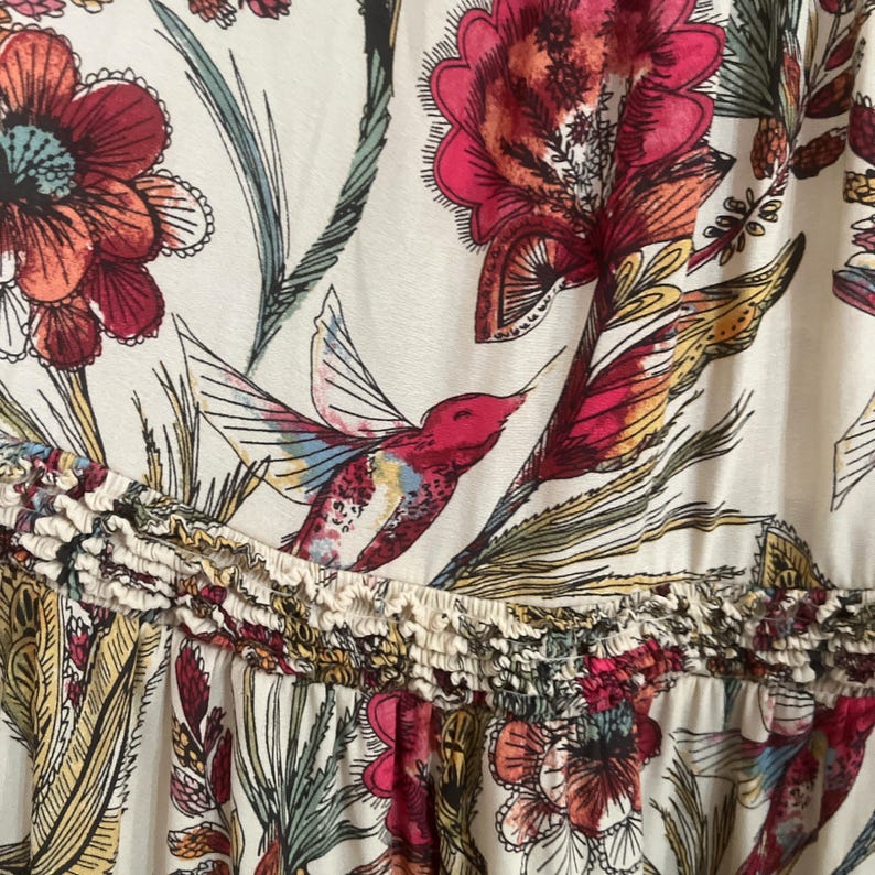 May include: Cream fabric with a colourful floral and hummingbird pattern. The design includes large red and pink flowers, green leaves, and a hummingbird with vibrant wings. A ruffled trim separates the top and bottom sections.