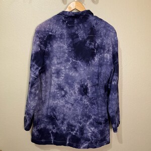 May include: A long-sleeved, button-down shirt with a tie-dye pattern in shades of blue and purple. The shirt is hanging on a wooden hanger against a neutral-colored wall. The tie-dye design creates a swirling, abstract effect.