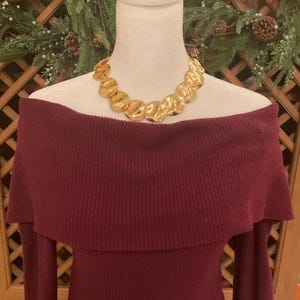 Puede incluir: Un vestido burdeos con hombros descubiertos y mangas largas se exhibe en un maniqu&iacute;. Un collar dorado con un dise&ntilde;o org&aacute;nico y texturizado adorna el escote. El fondo presenta un arreglo festivo de vegetaci&oacute;n y pi&ntilde;as.